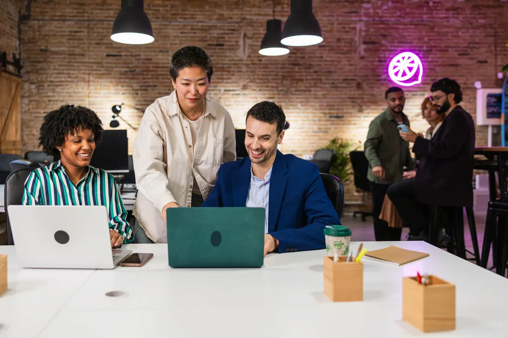 Drei Personen arbeiten gemeinsam an Laptops an einem Tisch in einem modernen, offenen Büro; zwei sitzen und lächeln, eine steht daneben und zeigt auf den Bildschirm, während im Hintergrund weitere Kolleg*innen miteinander sprechen.